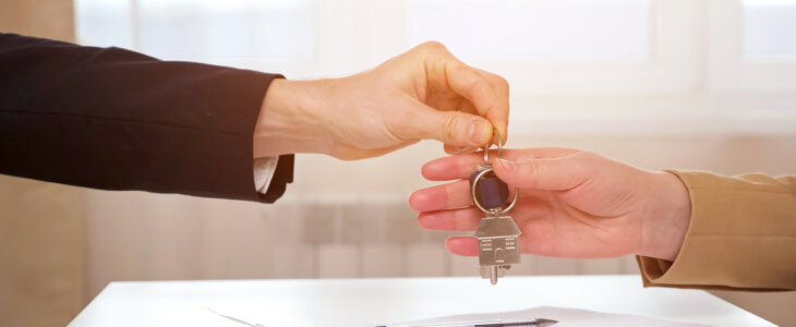 Real Estate Agent gives keys with house shaped breloque to young lady after signing of apartment rental contract at table in office, sunlight