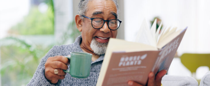 Elderly man, book and coffee on sofa with smile, reading or relax in retirement in home living room. Senior person, literature and happy with tea cup for knowledge, thinking and drink on lounge couch