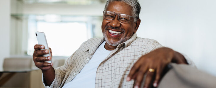 Cheerful man enjoying modern technology at home, sitting comfortably on the couch and smiling joyfully while holding a smartphone