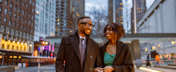 couple elegantly dressed for an evening out, walking in Manhattan, NYC. The city lights and tall buildings surround them as they warmly smile at each other.